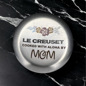 May include: A round, silver-colored Le Creuset lid with floral accents and the text "LE CREUSET COOKED WITH ALOHA BY MOM" in a handwritten font. The lid is set against a black and white marble background.