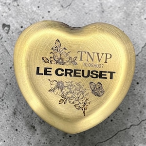 Op de afbeelding: Een hartvormig metalen deksel in goudtint met een geborstelde afwerking. Het oppervlak is gegraveerd met de tekst LE CREUSET, TNVP en de datum 10.06.2007, vergezeld van gedetailleerde zwarte lijntekeningen van bloemstengels en een vlinder.
