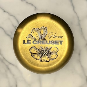 May include: Gold-colored metal lid with a floral design and the text "Le Creuset" and "Harvey" engraved on it.
