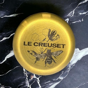 May include: A gold-coloured round dish with a black and white design of a bee and honeycomb. The text "LE CREUSET" is printed in black on the dish.