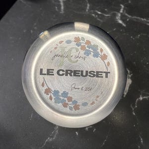 Peut inclure: Fond d'un gobelet Le Creuset en acier inoxydable avec texte gravé et motif floral. Le texte indique "janelle + chris", "LE CREUSET" et "June 8, 2024". Le motif floral comprend des accents bleus, oranges et verts.