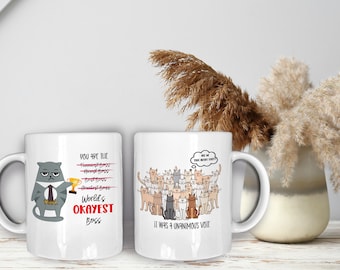 Op de afbeelding: Twee witte keramische mokken met humoristische kattenontwerpen. De eerste mok heeft een chagrijnige kat met een stropdas en een trofee in zijn hand, met de tekst "You are the World's Okayest Boss". De tweede mok toont een groep katten met een gedachteballon die zegt "But he gives good head scratches", en de tekst "It was a unanimous vote".