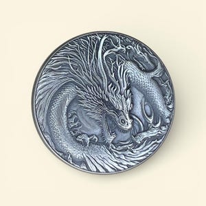 Op de afbeelding: Een gedetailleerde, cirkelvormige, zilverkleurige reliëfsculptuur van een draak. De draak is afgebeeld met ingewikkelde schubben, een vloeiende manen en een open mond, tegen een achtergrond van gestileerde bomen en water. Het kunstwerk is gemonteerd op een lichtbeige oppervlak.