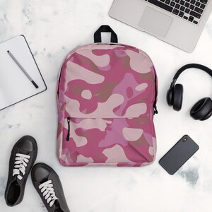 Puede incluir: Una mochila de camuflaje rosa con cremallera y asa negras. La mochila está rodeada por un portátil, auriculares, un smartphone, un cuaderno y zapatillas negras.
