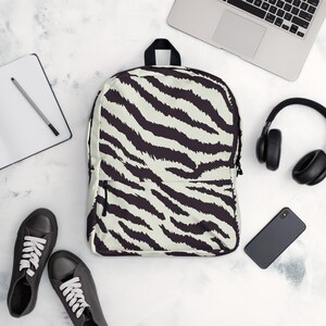 Könnte beinhalten: Ein Rucksack mit einem schwarz-weißen Zebrastreifenmuster. Der Rucksack hat einen schwarzen Reißverschluss und einen schwarzen Griff. Das Bild zeigt auch schwarze Turnschuhe, einen Laptop, Kopfhörer, ein Smartphone und einen Notizblock.