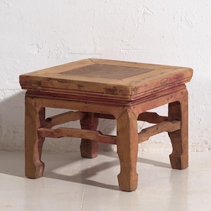 Può includere: Uno sgabello quadrato in legno invecchiato, di colore marrone rossiccio. Presenta un piano rialzato con un intarsio in legno più chiaro e gambe decorative angolate. Il design suggerisce uno stile antico o vintage.
