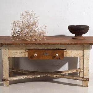Peut inclure: Une table en bois rustique avec un plateau marron et une base blanche vieillie. Elle comporte un seul tiroir avec des boutons décoratifs et un bol en bois foncé. Un arrangement floral séché repose sur la table.