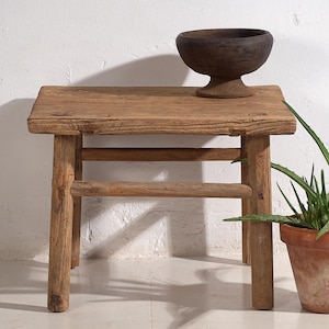 May include: A rustic wooden side table with a rectangular top and four sturdy legs. A dark brown wooden bowl sits on the table's surface. The table and bowl have a weathered appearance, suggesting age and use. A potted plant is in the background.