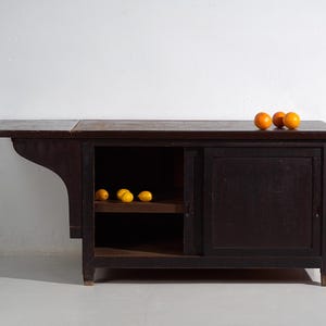 Peut inclure: Un meuble en bois brun foncé avec une table pliante. Le meuble a une porte coulissante et une étagère ouverte. Trois oranges sont posées dessus et plusieurs citrons sont à l'intérieur. Le meuble est contre un mur blanc.