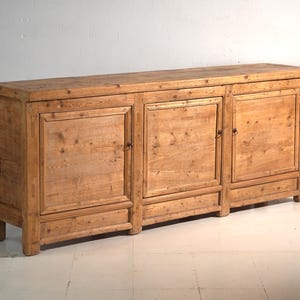 Op de afbeelding: Een lang, rechthoekig houten dressoir met een natuurlijke lichtbruine afwerking en zichtbare houtnerf. Het meubelstuk heeft drie kastdeuren, elk met een eenvoudig kaderontwerp en kleine ronde metalen handgrepen, rustend op vier stevige blokpoten.