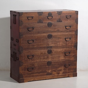 Peut inclure: Une commode en bois brun foncé avec cinq tiroirs. Le coffre a du matériel en métal sur les coins et autour des poignées de tiroir. Les poignées de tiroir sont en forme de D. Le coffre est de style traditionnel.