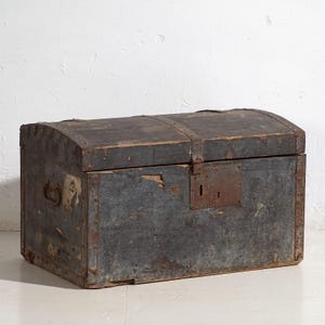 Könnte beinhalten: Ein verwitterter, antiker Holzkoffer mit gewölbtem Deckel und Metallbeschlägen. Der Koffer ist in einem verblichenen Blau-Grau gestrichen und weist Gebrauchsspuren auf. Mit einem rostigen Metallschloss und Griff. Eine Vintage-Truhe.