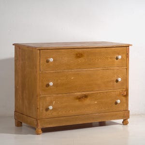 May include: A three-drawer wooden chest of drawers with a light brown finish. It features white, round knobs and rests on short, rounded legs. The wood grain is visible across the drawers and top.