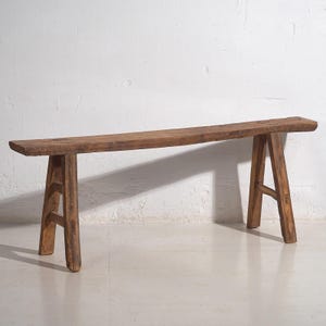 Può includere: Panca rustica in legno con seduta lunga e rettangolare e gambe a forma di A. La panca è realizzata in legno stagionato, che mostra venature e nodi naturali. La panca è di colore marrone caldo e sembra fatta a mano.