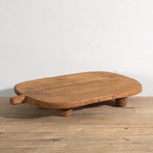 May include: A wooden serving board with a handle and three small feet. The board is made of light brown wood and has a natural, unfinished appearance. The handle is on the left, and the feet are evenly spaced. The board is approximately 40 cm long.