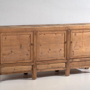 Könnte beinhalten: Ein großer, rechteckiger Holzschrank mit drei Türen und einer Schublade. Der Schrank ist aus hellbraunem Holz gefertigt und hat ein rustikales, gealtertes Aussehen. Der Schrank wird von vier kurzen Beinen getragen.