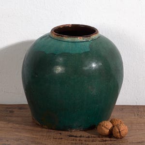 Op de afbeelding: Een grote, ronde, antieke groene keramische pot met een donkerbruin interieur en rand. De pot heeft een glanzende afwerking en staat op een houten oppervlak. Drie walnoten liggen naast de pot.