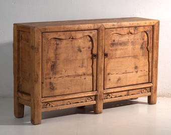 May include: A rectangular wooden sideboard cabinet featuring a natural light brown finish with visible wood grain and knots. The piece has two doors with arched top panels, small circular metal pulls, and decorative carved trim along the base.