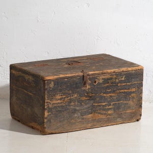 Könnte beinhalten: Eine rechteckige Holzkiste mit abgenutztem Aussehen. Die Kiste ist in dunklen Grau- und Brauntönen lackiert, mit sichtbarer Holzmaserung und alter Metallbeschlägen. Der Deckel ist geschlossen und mit einem Riegel gesichert.
