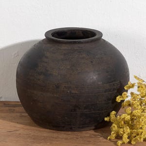 Op de afbeelding: Een donkerbruine, ronde keramische vaas met een brede opening. De vaas heeft horizontale lijnen rondom de body en staat op een houten oppervlak. Gele bloemen bevinden zich rechts van de vaas.
