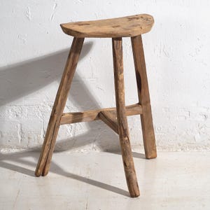 Può includere: Sgabello rustico in legno con seduta curva e tre robuste gambe. Lo sgabello è realizzato in legno marrone chiaro e ha un aspetto naturale e non rifinito. Le gambe sono angolate per la stabilità e il design è semplice e funzionale.
