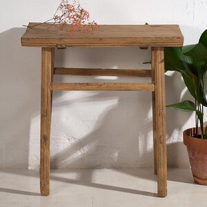 May include: A weathered, rectangular wooden side table with four legs. The table has a natural wood grain finish and a simple, rustic design. A small sprig of dried orange flowers rests on the tabletop. The table is approximately 76 cm long.