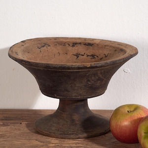 Può includere: Una fruttiera rustica in legno marrone scuro, con una base arrotondata e un ampio bacino poco profondo. Due mele rosse e verdi sono poste accanto alla ciotola su una superficie di legno.
