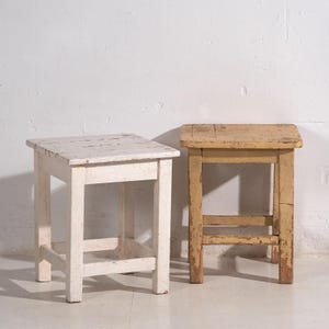 Op de afbeelding: Twee kleine, vierkante houten krukken. Eén is wit geschilderd en vertoont tekenen van slijtage. De andere is lichtgeel met zichtbare houtnerf en afbladderende verf. Beide hebben vier poten en een eenvoudig, rustiek ontwerp.