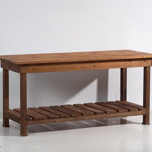 Op de afbeelding: Een rechthoekige houten tafel met een onderste plank van latten. De tafel is gemaakt van bruin hout en heeft een eenvoudig, stevig ontwerp. De tafel is ongeveer 152 cm lang.