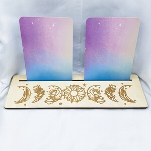 Op de afbeelding: Een houten kaarthouder met een maan- en zonontwerp. De houder heeft twee sleuven voor kaarten en is gemaakt van lichtbruin hout. Het ontwerp is in het hout gegraveerd en bevat een halve maan, een volle maan, een zon en bloemen.