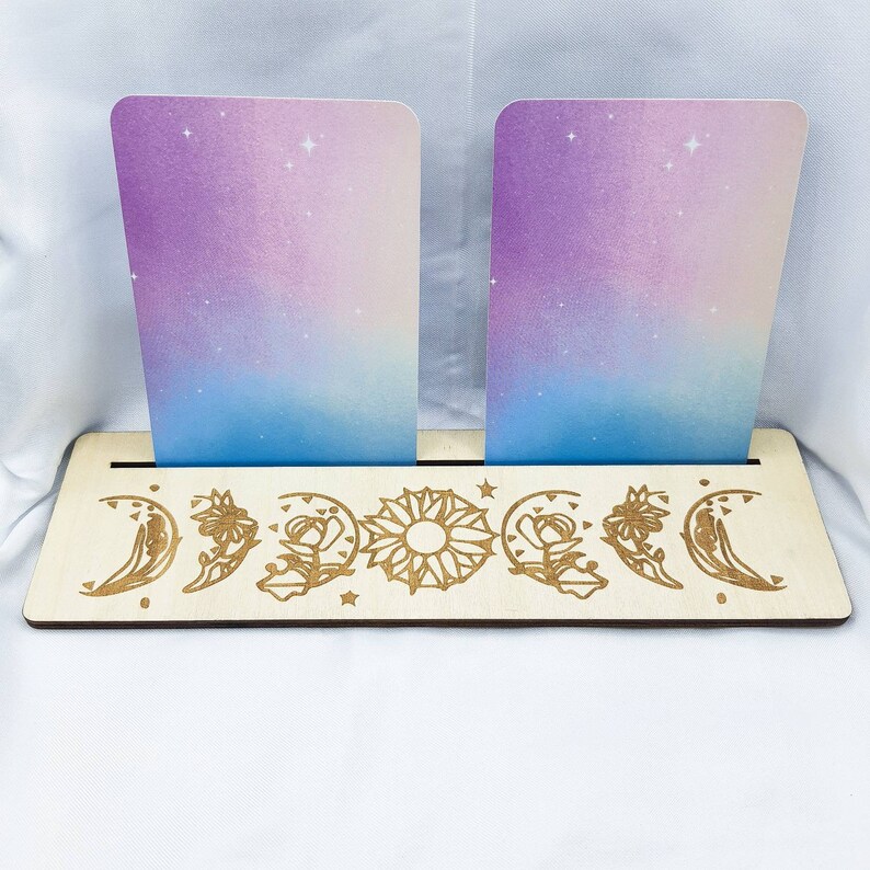 Op de afbeelding: Een houten kaarthouder met een maan- en zonontwerp. De houder heeft twee sleuven voor kaarten en is gemaakt van lichtbruin hout. Het ontwerp is in het hout gegraveerd en bevat een halve maan, een volle maan, een zon en bloemen.