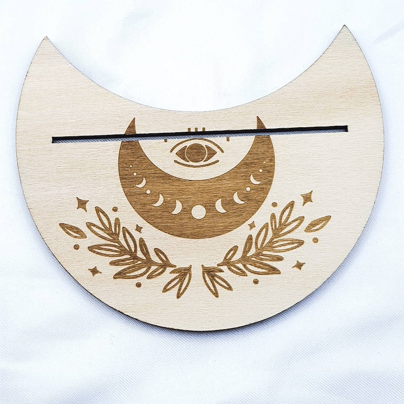 Op de afbeelding: Een houten halve maan wierookhouder met een gedetailleerd ontwerp van een maan met fasen, een oog en bladeren. De houder heeft een gleuf voor wierookstokjes.