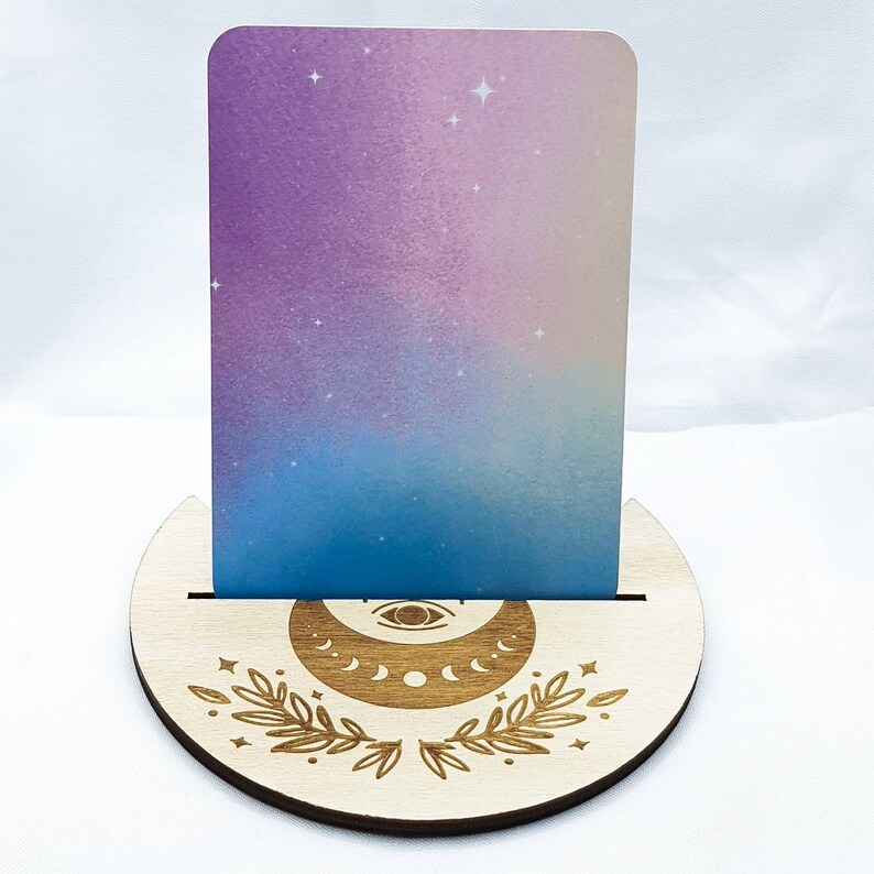 Op de afbeelding: Een houten kaarthouder met een halve maan en oogontwerp. De houder is gemaakt van lichtbruin hout en heeft een gleuf om een kaart vast te houden. De kaart in de houder is een lichtblauwe en paarse gradi&euml;nt met witte sterren.