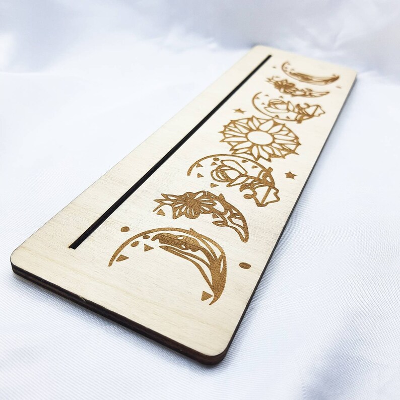 Op de afbeelding: Een houten bord met een zwarte lijn in het midden en een reeks maanfasen en een zon die rechts gegraveerd zijn. De maanfasen zijn gestileerd met bloemmotieven.