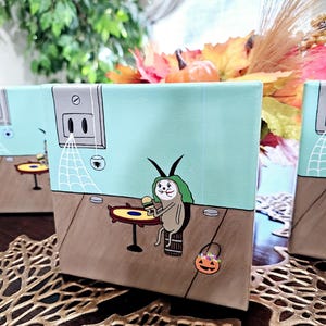May include: A hand-painted canvas featuring a cartoon bug wearing a green hat and holding a burger. The bug is sitting at a table with a spiderweb and a small pumpkin on the floor. The background is a light blue wall with a white electrical outlet.