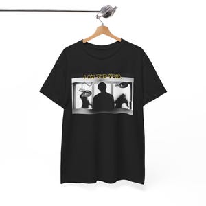 Może przedstawiać: Czarny t-shirt z grafiką przedstawiającą osobę w pokoju z dwiema cienistymi postaciami. Tekst "I AM THE VOID" jest wydrukowany nad grafiką w kolorze żółtym.