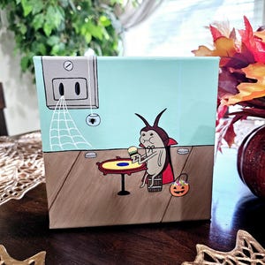 May include: A hand-painted canvas featuring a cartoon cockroach dressed as a vampire, holding a candy bar and sitting at a table with a spiderweb and a spider in the background. The cockroach is wearing a red cape and a black hat. The canvas is a light blue color with brown accents.