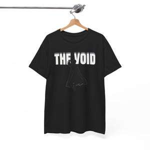 May include: A black short-sleeve t-shirt featuring the white text THE VOID printed across the chest. Below the text is a white line-art graphic of a figure wearing a long flowing cape and reaching forward with one hand, centered on the shirt.