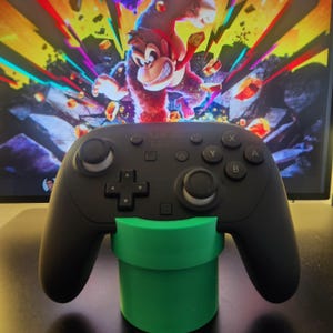 May include: A black video game controller resting on a green pipe-shaped stand. The controller has buttons labeled A, B, X, and Y. In the background, a colorful video game scene featuring a cartoon character is visible.