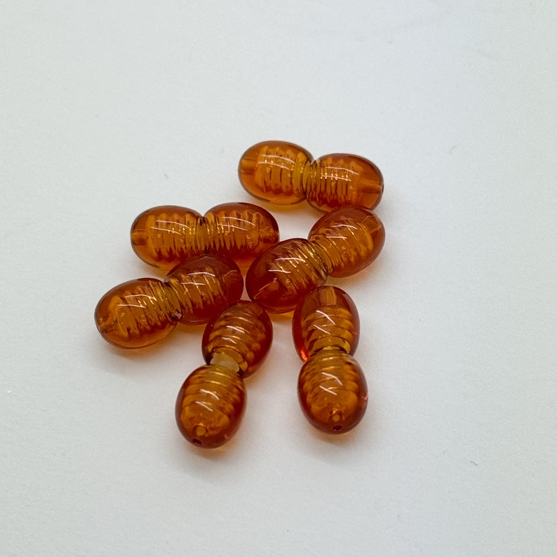May include: A group of six amber-colored glass beads, each with a twisted design. The beads are arranged in a loose pile on a white surface.