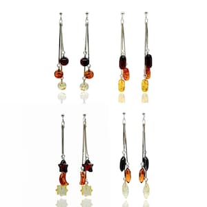 May include: Four pairs of dangle earrings with silver-tone metal posts and long, slender bars. Each earring features a unique arrangement of amber-coloured beads and charms, including round, oval, and star shapes. The amber hues range from light yellow to deep brown.