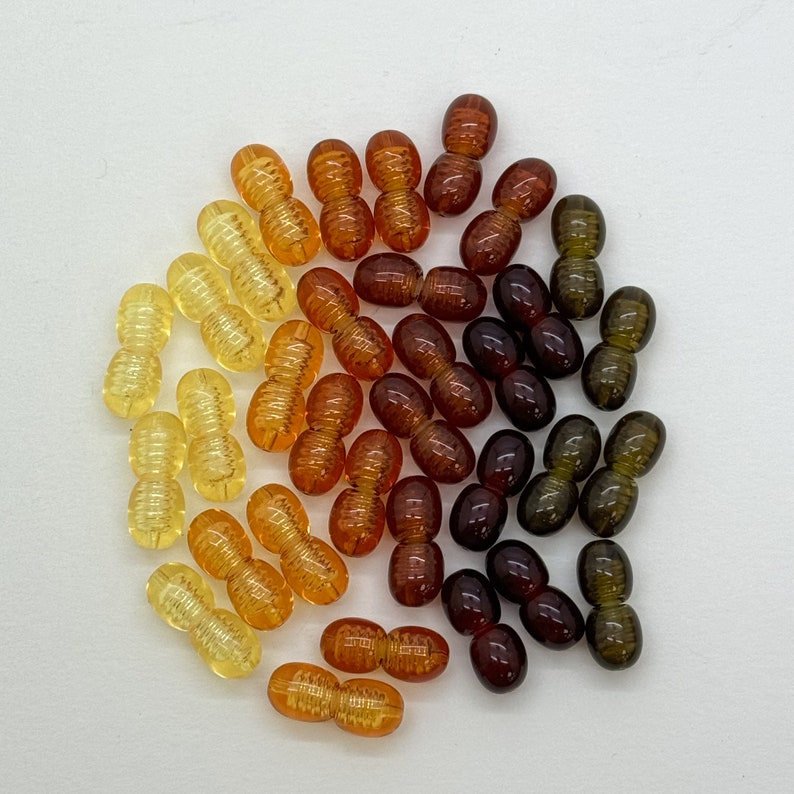 May include: A collection of amber, brown, and green glass beads. The beads are all in the shape of a peanut and are arranged in a circle.