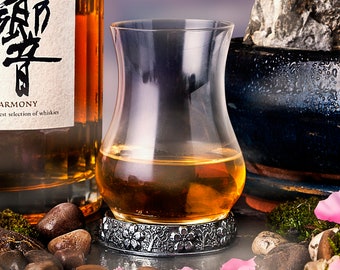 Pode incluir: Uma garrafa de whisky japonês Hibiki Harmony com um copo de whisky à sua frente. A garrafa é transparente e tem uma superfície texturizada. O copo é transparente e tem uma base redonda. Há uma árvore bonsai no fundo. O fundo é uma imagem desfocada de uma cordilheira.