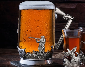 May include: A set of three silver-plated shot glasses with a hunting scene design. The glasses are filled with a dark amber liquid and are sitting on a wooden table with shotgun shells. The glasses are surrounded by a burlap cloth.