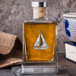 May include: A glass decanter with a silver sailboat emblem, filled with amber liquid. The decanter sits on a silver base with the text "A E WILLIAMS MADE IN UK". It has a cork stopper and silver cap. The background includes a burlap sack and a blue and white bowl.