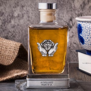 Peut inclure: Un flacon en verre transparent rempli d'un liquide ambré, orné d'un emblème argenté de mains tenant un motif. Le flacon repose sur une base en ardoise, avec le texte "A E WILLIAMS MADE IN UK". Un sac en toile de jute est en arrière-plan.