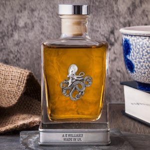 Könnte beinhalten: Eine klare Glasflasche mit bernsteinfarbener Flüssigkeit, verziert mit einem silbernen Tintenfisch-Emblem. Sie hat einen silbernen Verschluss und steht auf einem Schieferfuß. Der Text "A E WILLIAMS MADE IN UK" ist auf dem Sockel zu sehen.