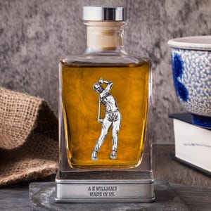 May include: A clear, square-shaped decanter filled with amber liquid, featuring a silver golfer figure. The decanter has a silver top and a wooden cork. The base reads "A E WILLIAMS MADE IN UK".
