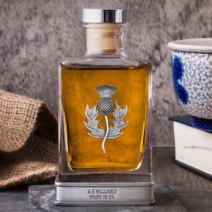 May include: A square glass decanter filled with amber liquid, adorned with a silver thistle emblem. The decanter has a silver stopper and sits on a silver base with the text "A E WILLIAMS MADE IN UK".