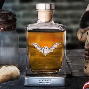 May include: A square glass bottle with a silver bat emblem, filled with amber liquid, sits on a metal stand. The bottle is labeled "A E Williams Made in UK." Other antique bottles, bones, and a skull are in the background.